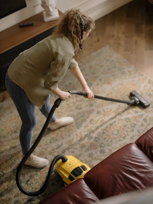 A person using a yellow and black vacuum cleaner for surface cleaning on a patterned carpet in a domestic setting. The individual is kneeling on the carpet, reaching into the vacuum's open dust compar