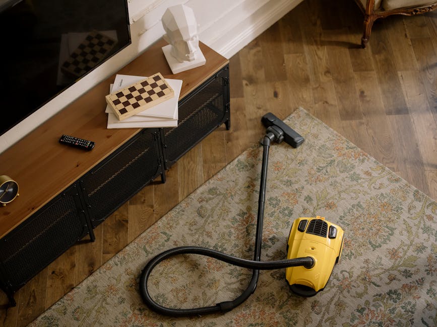 A person dressed in full protective cleaning gear, including a white suit, gloves, and blue shoe covers, is using a vacuum or carpet cleaning machine on a beige, textured carpet in a bright, modern li