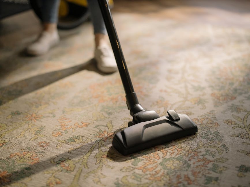 A person using a handheld vacuum cleaner to deep clean a patterned area rug in a domestic setting. The individual's lower legs and white sneakers are visible, with the vacuum hose extending from a yel