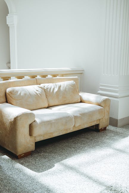 A white upholstered sofa with tufted backrest and rolled arms, positioned against a white wall with decorative molding in a well-lit room. The sofa has a clean appearance with no visible dust or stain